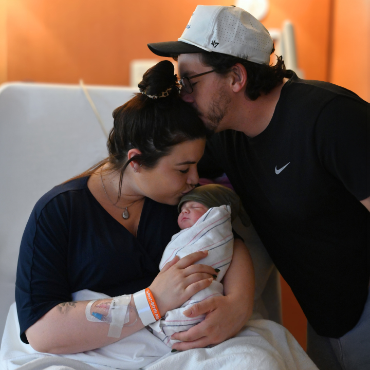 A haooy couple embraces with their newborn, while momwears an orange post-partum bracelet