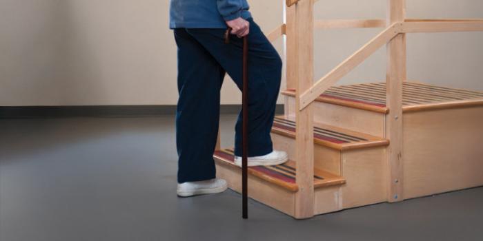 A person's hand grips onto the railing as they step up stairs one leg at a time - using the non-operated leg up first, followed by the operated leg.